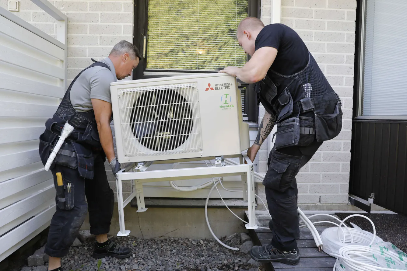 Kaksi asentajaa asentavat Mitsubishi Electric -ilmastointilaitetta valkoiselle jalustalle ulkona, valkoisen seinän ja ikkunan edessä.